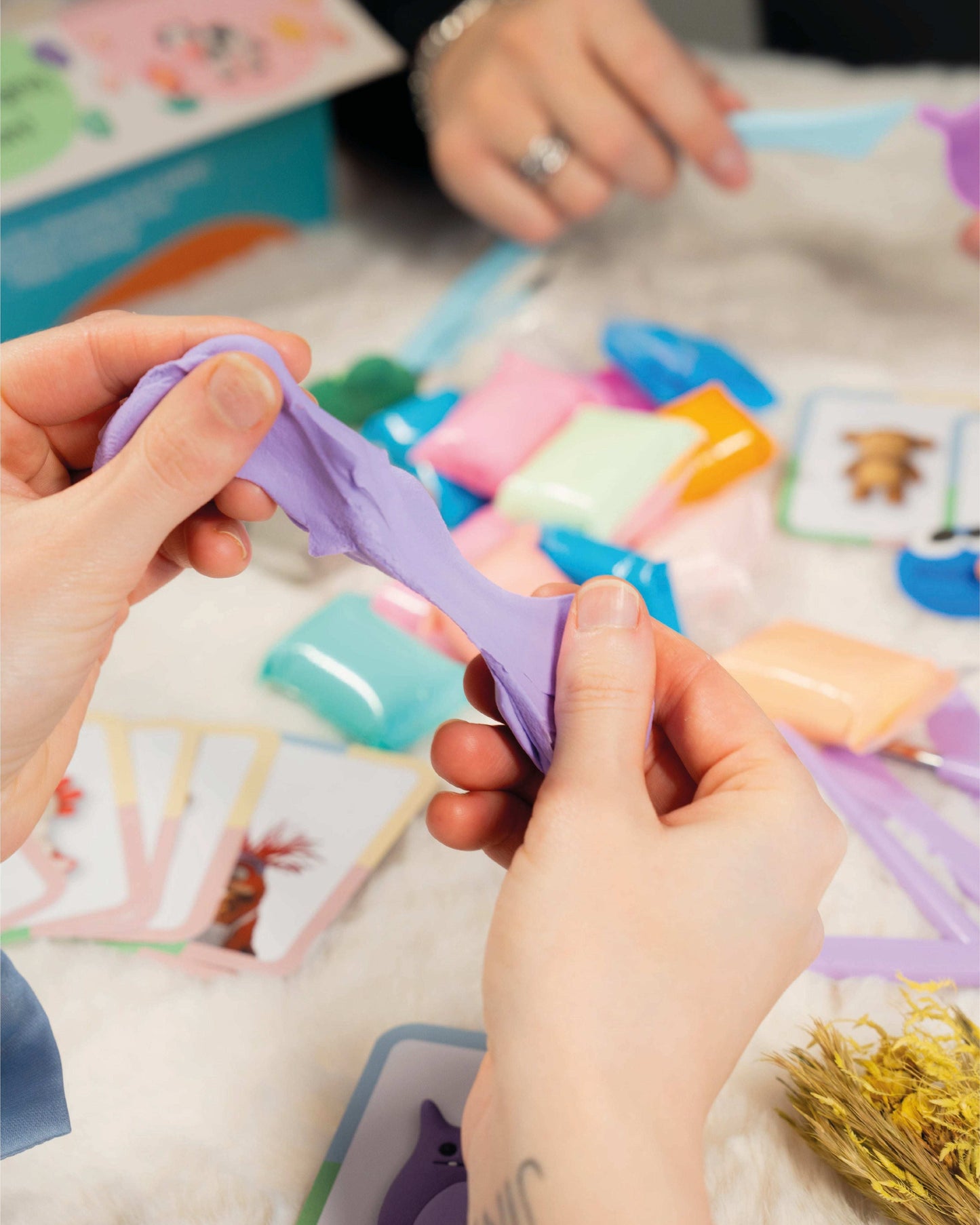 Hands holding a purple plastic tool over a colorful assortment of toys on a Clay Date™ Kit, with Extra Clay Pack