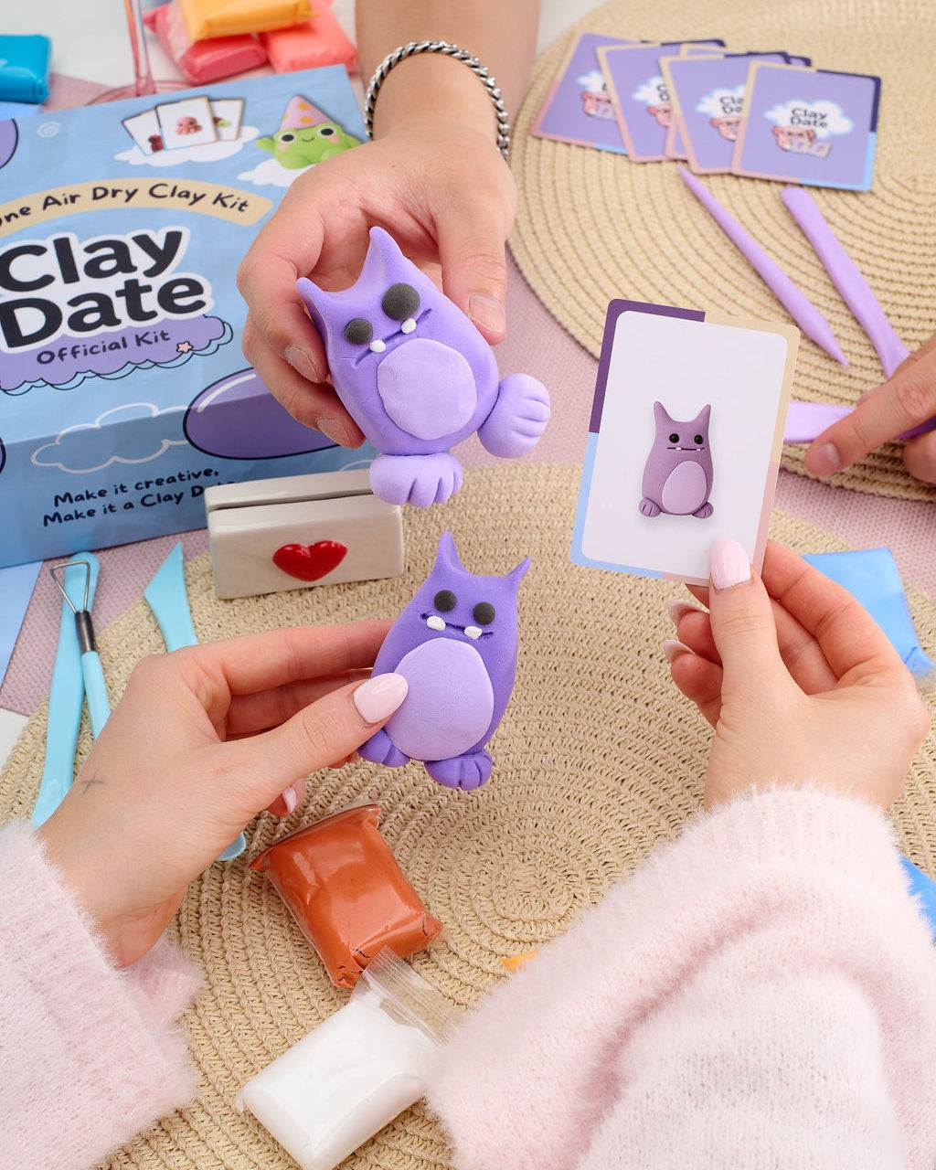 A person's hands holding purple clay figurines and an owl-themed card, with an air dry clay kit box and crafting tools.