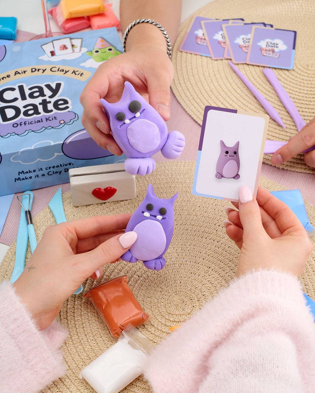 A person's hands holding purple clay figurines and an owl-themed card, with an air dry clay kit box and crafting tools on a woven mat background.