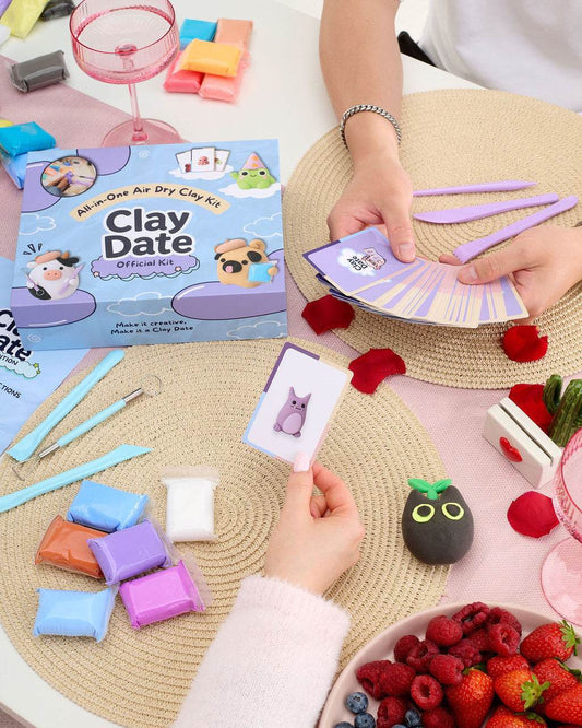 A person's hands are shown playing with a Clay Date activity kit, holding a card with a purple cat illustration and purple clay tools.