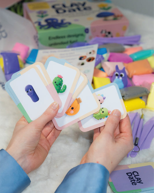 Hands holding colorful clay modeling cards with various shapes and colors, surrounded by clay and packaging.