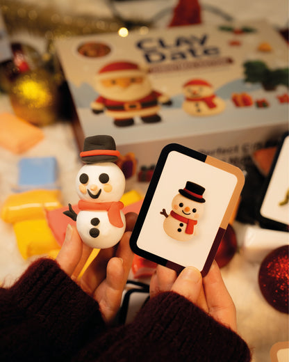 Hand holding a snowman figurine next to a card with a snowman design, with 'Clay Date' product in the background.