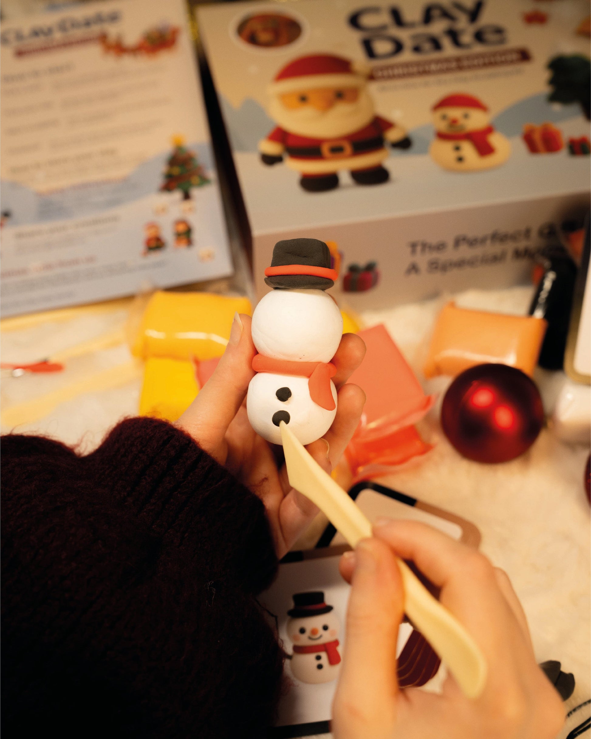 Person holding a small snowman sculpture made from clay with 'Clay Date' packaging in the background.