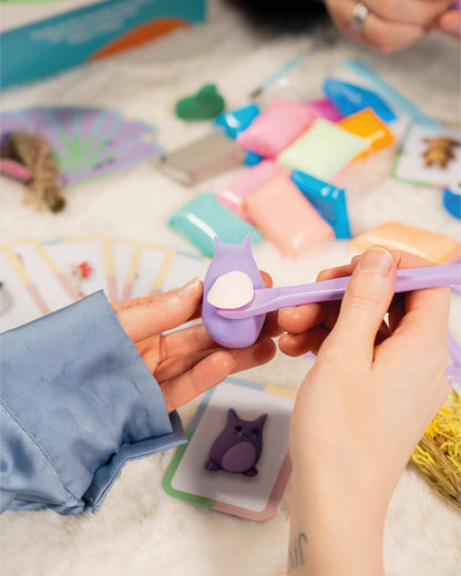 Person holding a small purple tool over a Clay Date™ Kit with cards and toys in the background.