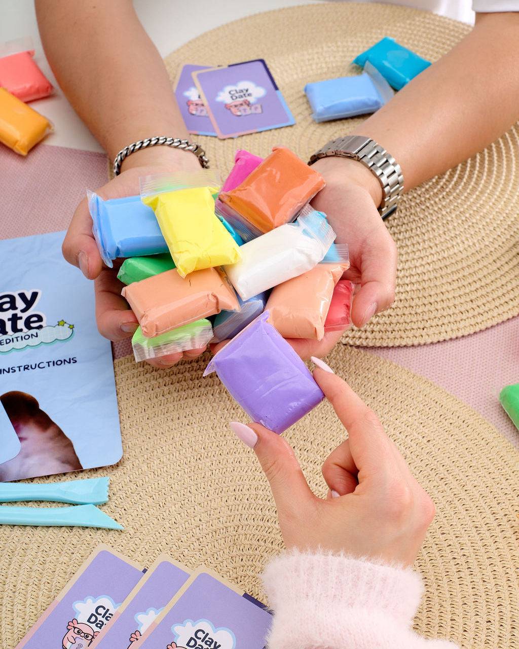 A person holds a handful of colorful clay pieces over their hands, with instruction cards and additional clay pieces arranged on woven mats and a table.