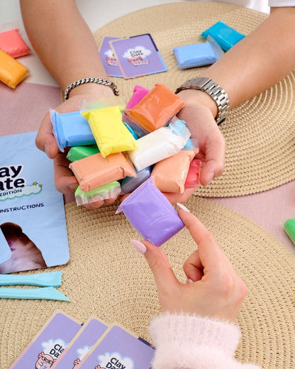A person holds a handful of colorful clay pieces over their hands, with instruction cards and additional clay pieces arranged on woven mats and a table.