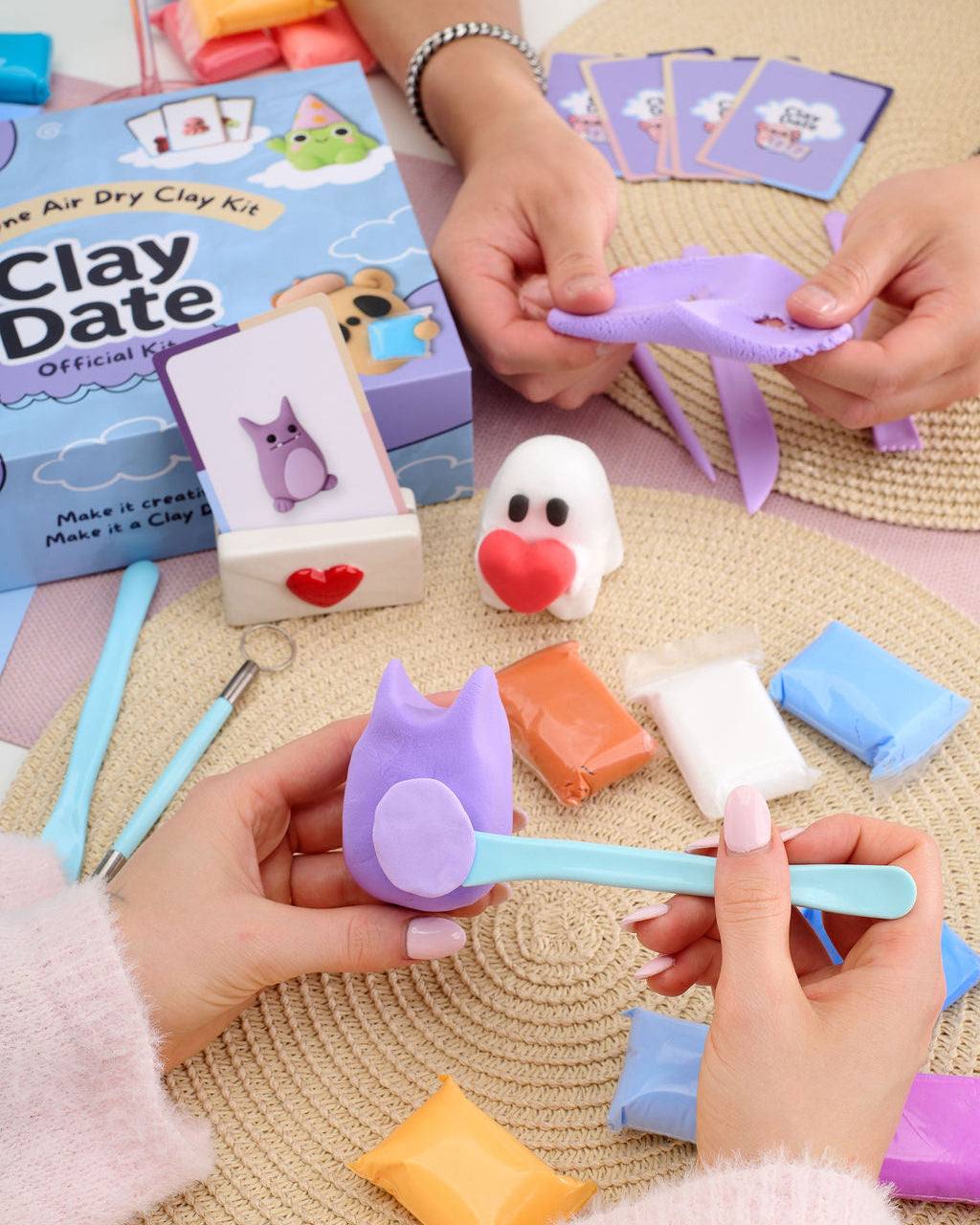 A person's hands are shown using a light blue air dry clay tool to shape a purple cat-shaped piece of clay, with a blue 'Clay Date' kit box and other colorful clay pieces on a woven mat.