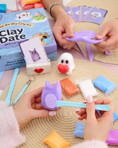 A person's hands are shown using a light blue air dry clay tool to shape a purple cat-shaped piece of clay, with a blue 'Clay Date' kit box and other colorful clay pieces on a woven mat.