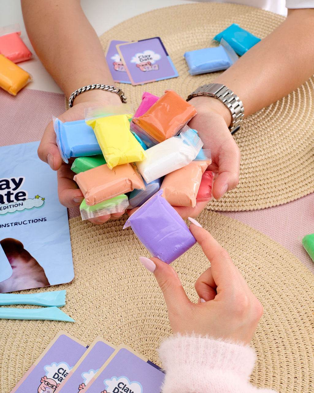 A person holds a handful of colorful clay pieces over their hands, with instruction cards and additional clay pieces arranged on woven mats and a table.