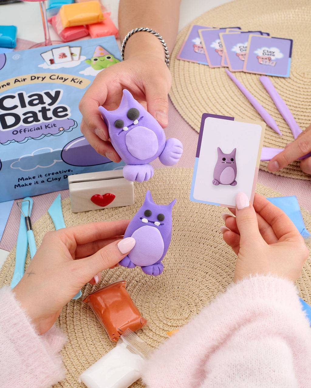 A person's hands holding purple clay owl figurines and an owl-themed card, with an air dry clay kit box and crafting tools on a woven mat background.