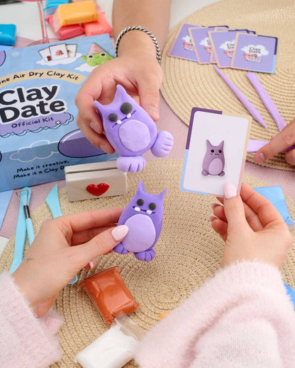 A person's hands holding purple clay owl figurines and an owl-themed card, with an air dry clay kit box and crafting tools on a woven mat background.