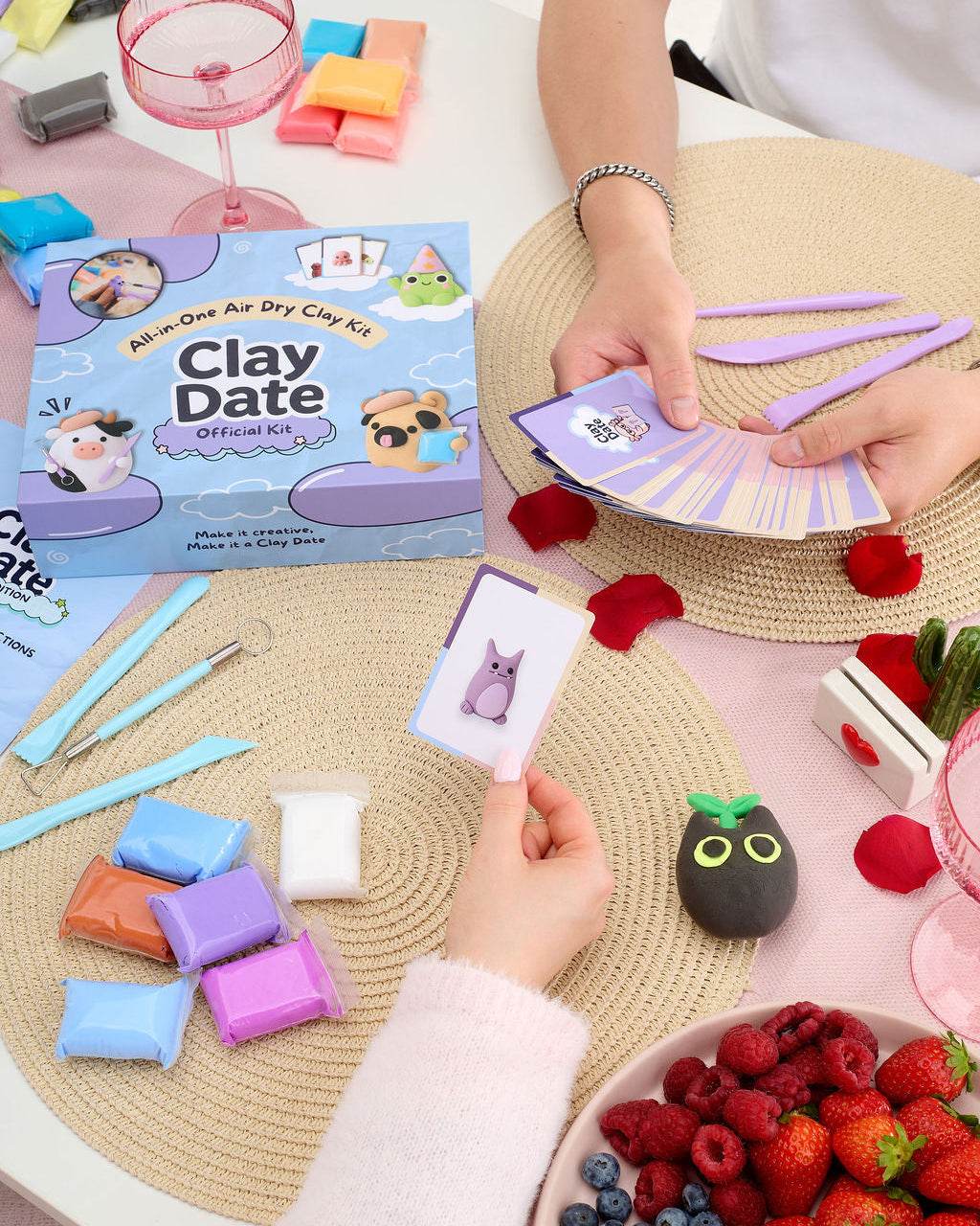 A person's hands are shown playing with a Clay Date activity kit, holding a card with a purple figure illustration and purple clay tools. 