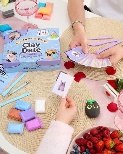 A person's hands are shown playing with a Clay Date activity kit, holding a card with a purple figure illustration and purple clay tools. 