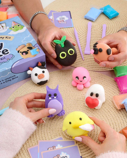 A colorful clay toy kit displayed on a woven beige mat, with hands holding various cute squishy clay characters.