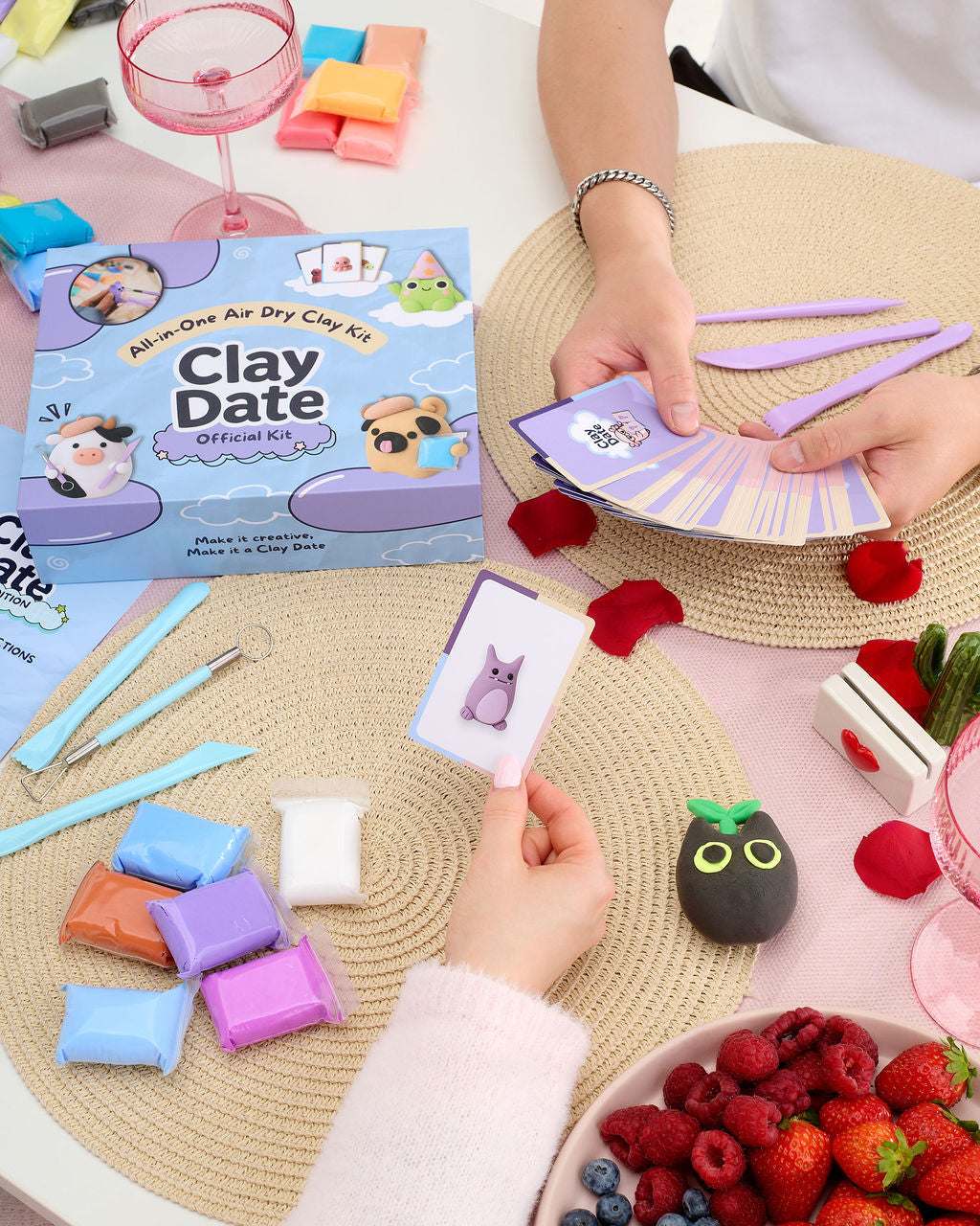 A person's hands are shown playing with a Clay Date activity kit, holding a card with a purple cat illustration and purple clay tools.