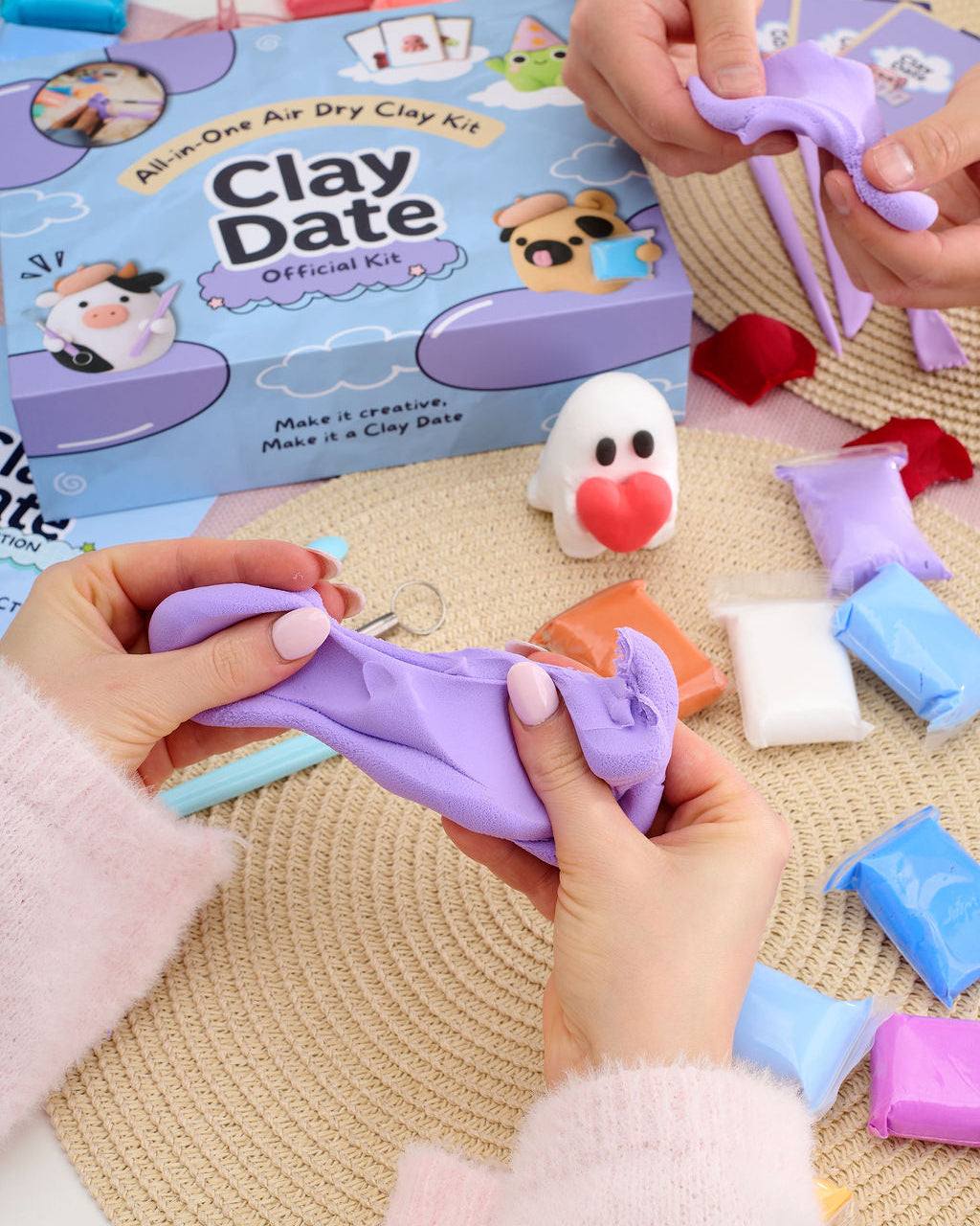 A person's hands stretching a purple clay figure over a woven beige mat, with a blue and purple 'Clay Date Official Kit' box in the background.